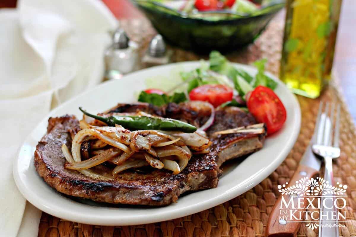 t-bone steak on a white plate with salad and topped with onions and peppers. 