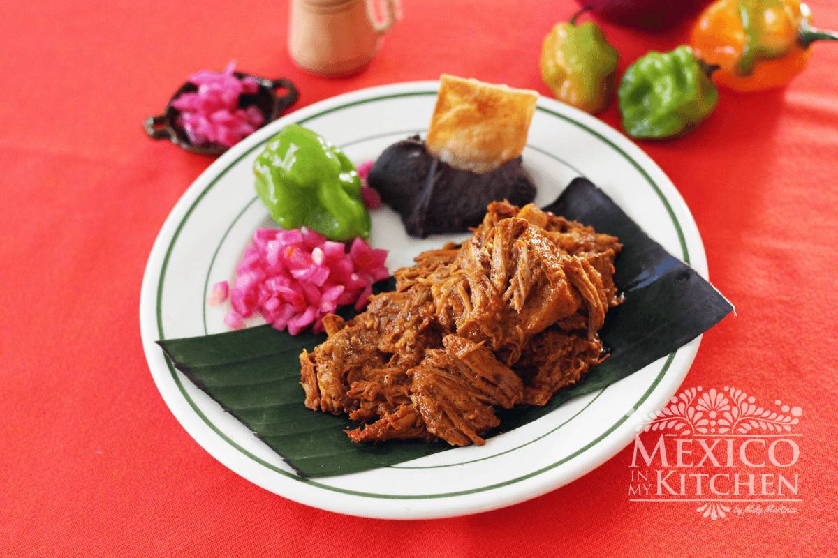 Pork cochinita, black beans, and pickled onions served on a banana leaf on top of a plate.