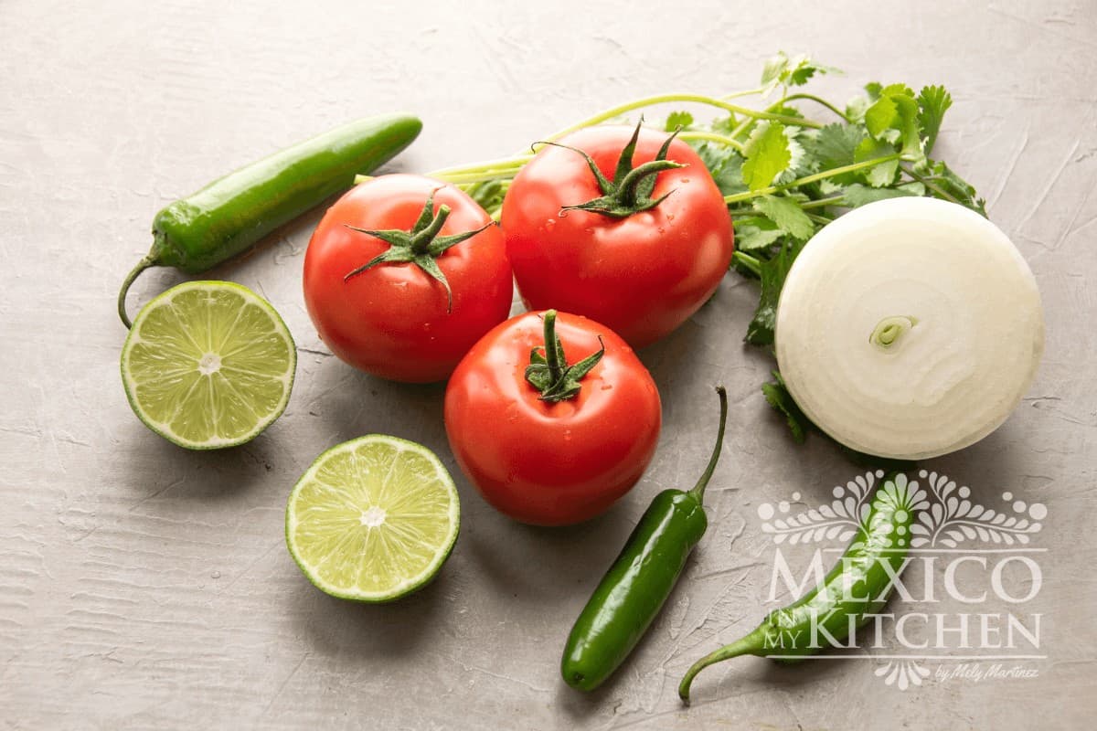 Tomatoes, onions, serrano peppers, cilantro and limes displayed on a board.