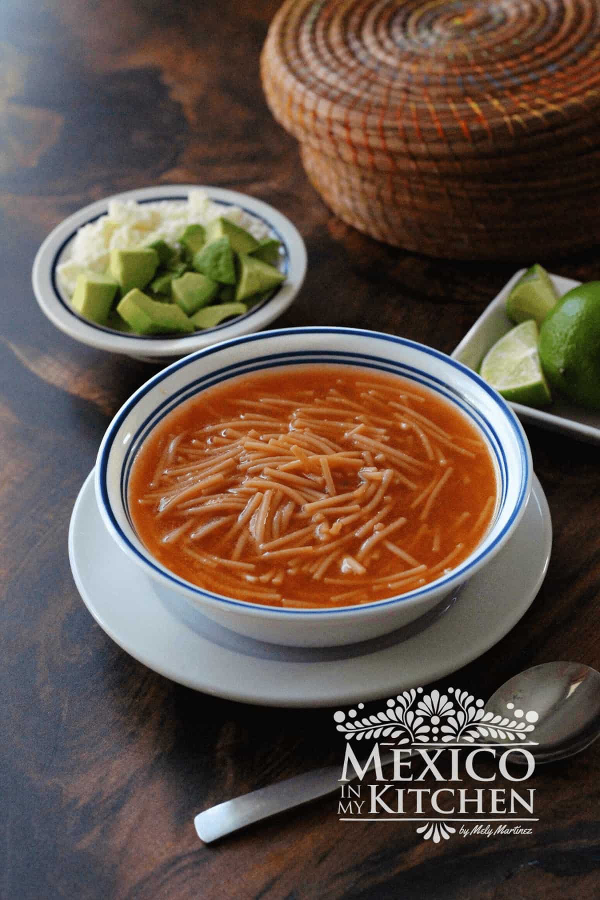 Sopa de fideo served in a bowl, next to limes and avocado.