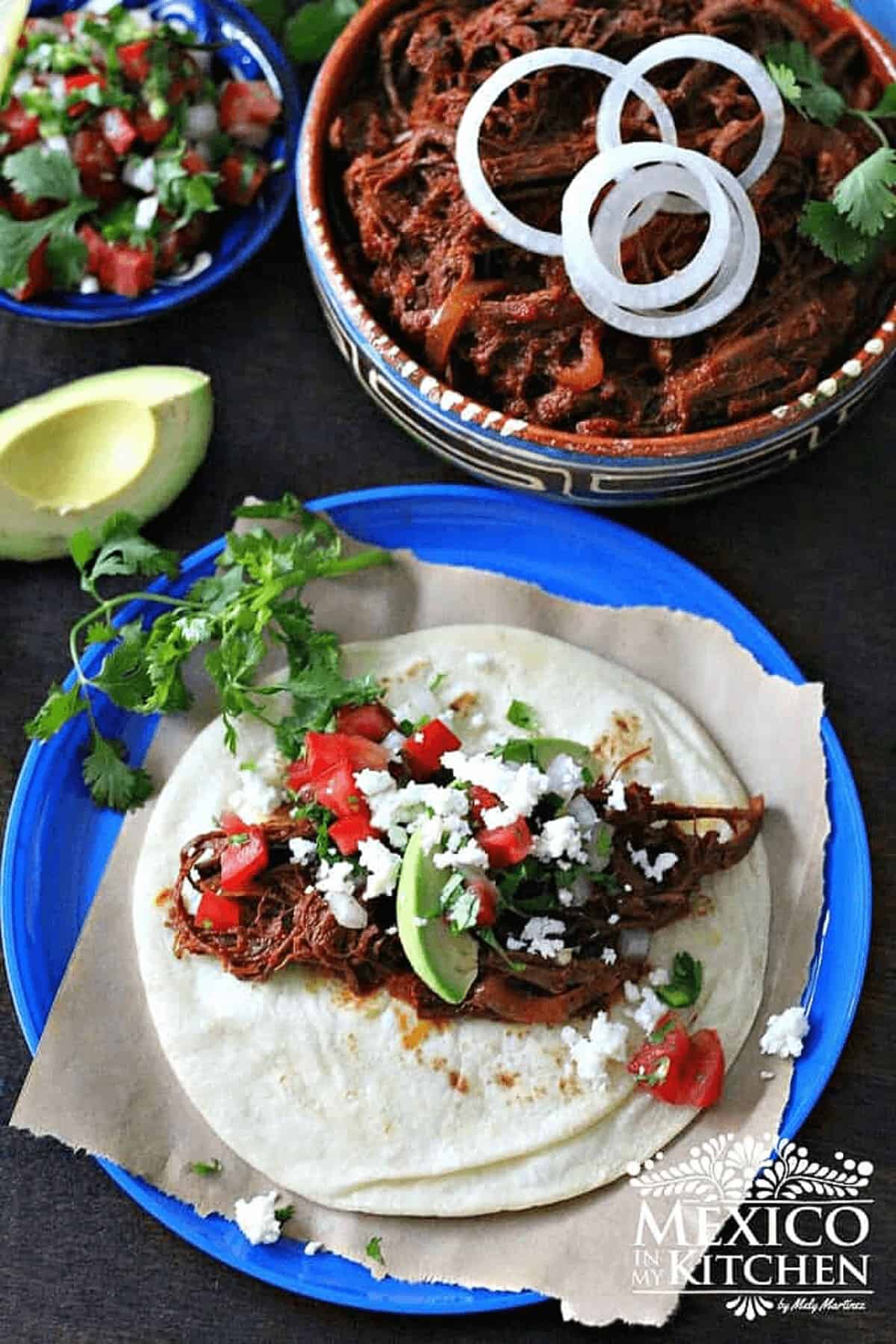Shredded beef with ancho chiles tacos, garnished with avocado, queso fresco and cilantro.
