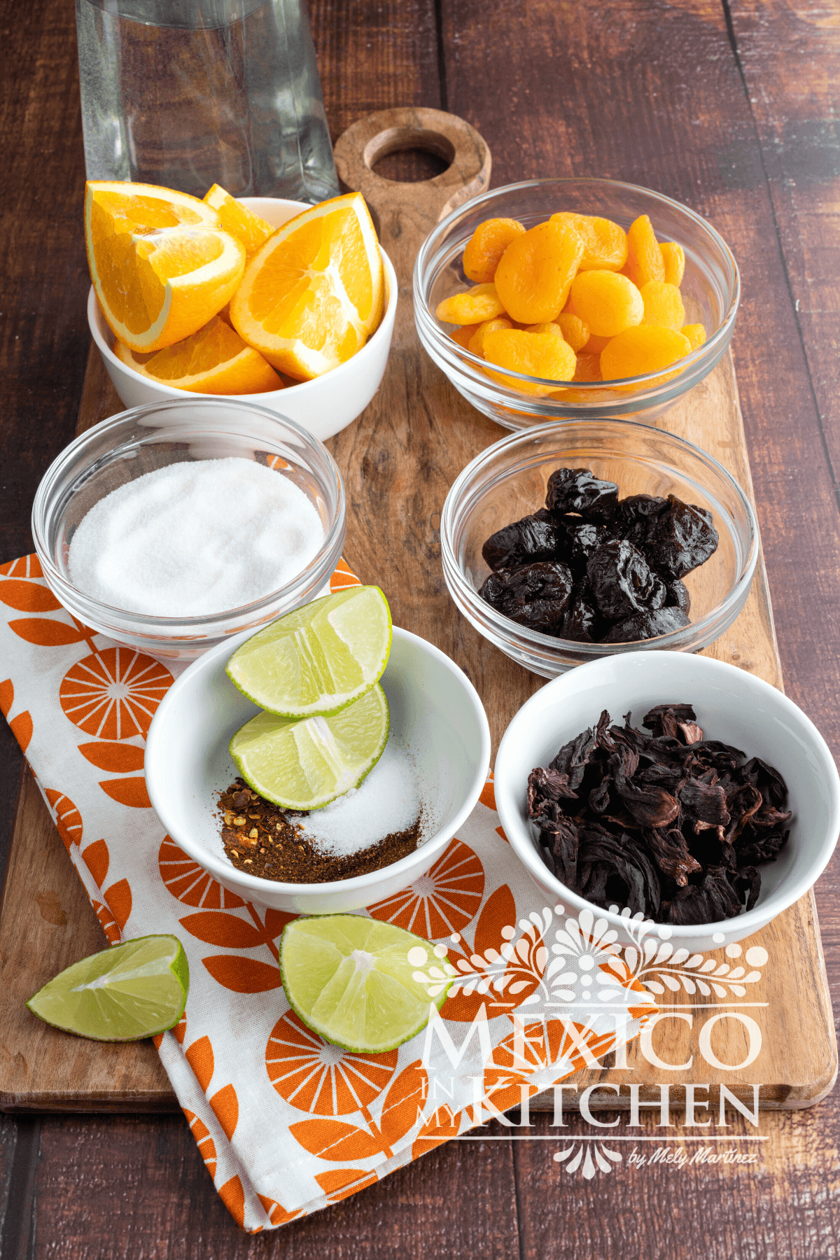 Ingredientes like oranges, dried dates, dried apricots, hibiscus flowers, limes, sugar and spices displayed in a table.