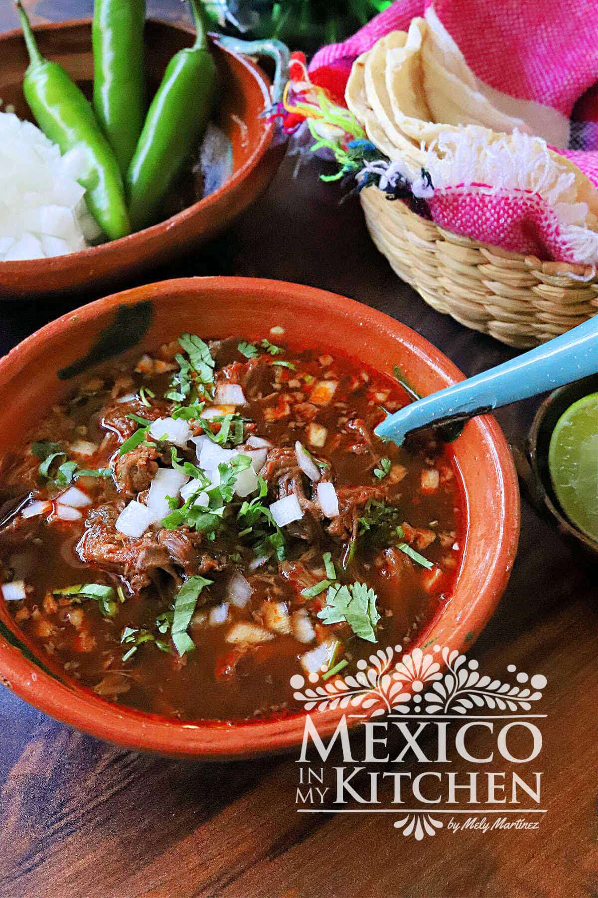 Beef Birria served in a red clay bowl and topped with fresh cilantro, and white onion.
