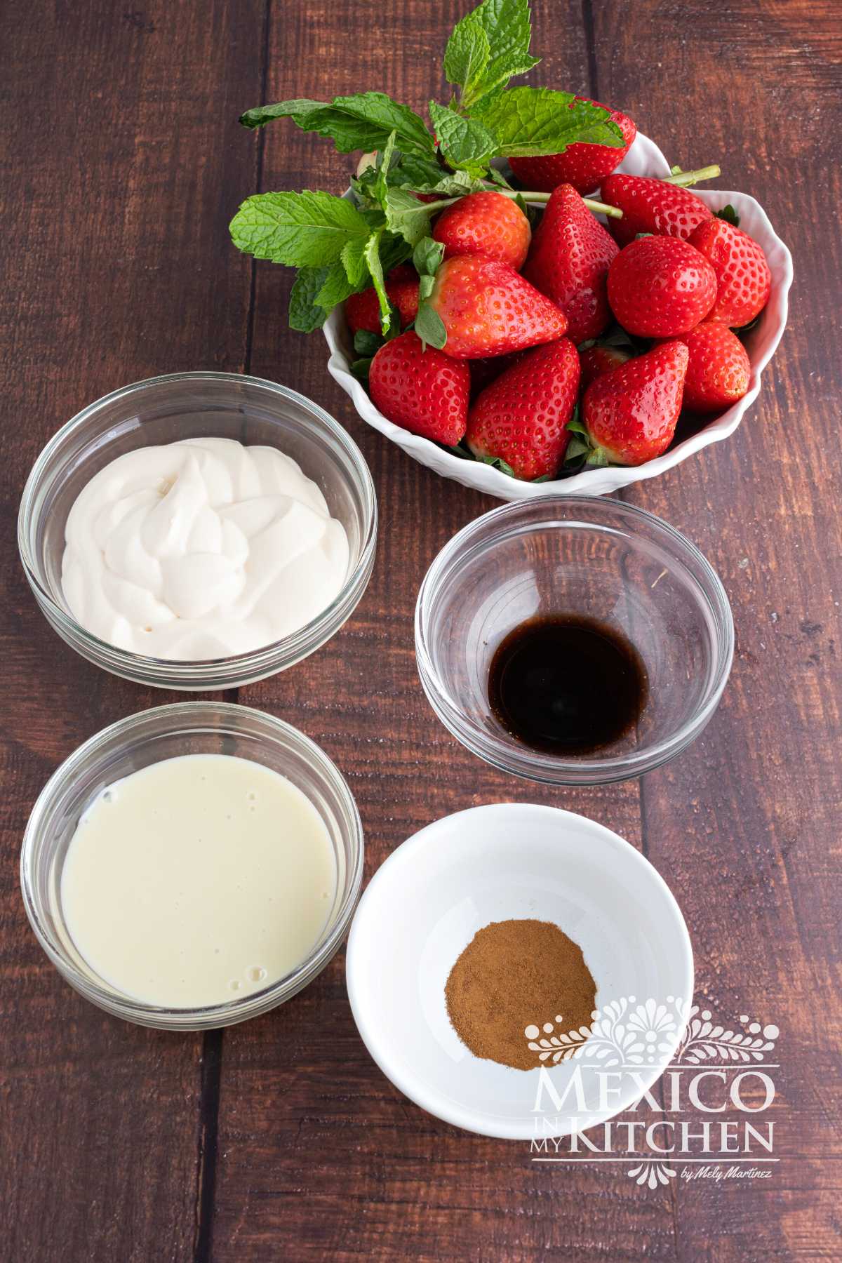 Ingredients like Mexican crema, condensed milk, vanilla, powder cinnamon, and strawberries are displayed on a table.