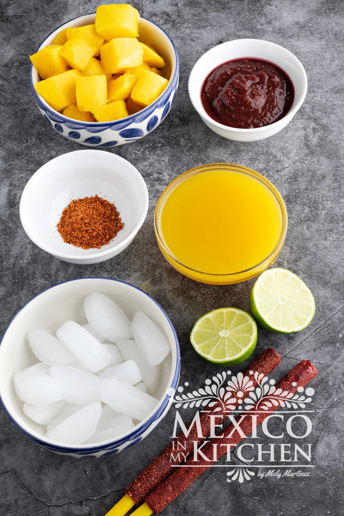 Ingredients like mango chunks, ice, mango nectar, Chamoy Sauce, tajin powder, limes are displayed on a countertop.