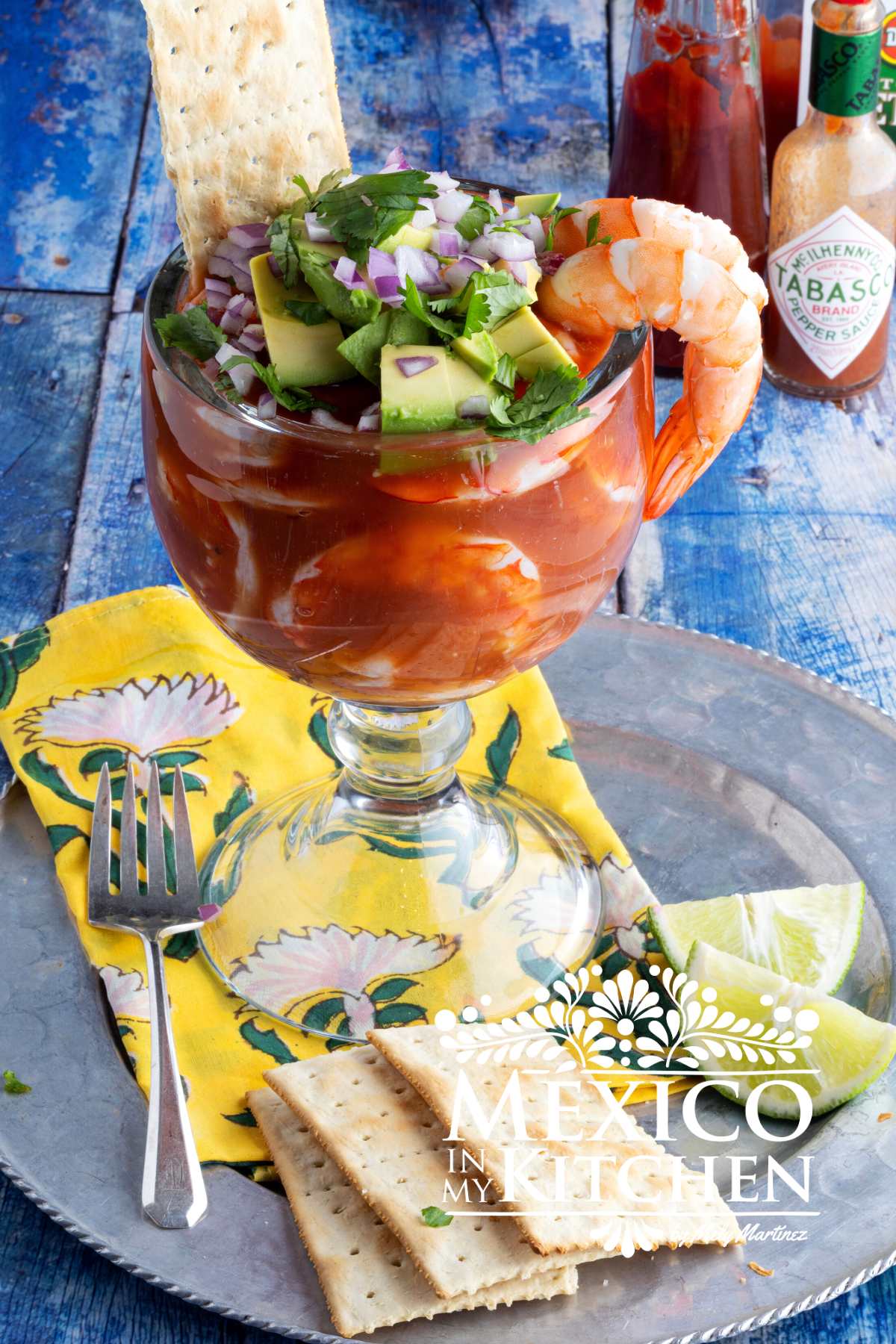 Mexican Shrimp cocktail served in a big glass bowl topped with avocado, cilantro and chopped purple onion next to saltines and lemons.