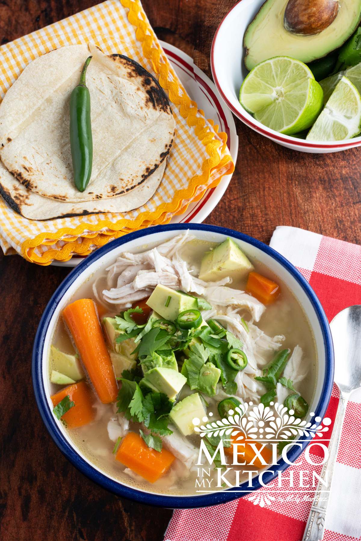 Caldo de Pollo (Mexican chicken soup) served in a white bowl, next to another bowl with avocado, cilantro and limes.