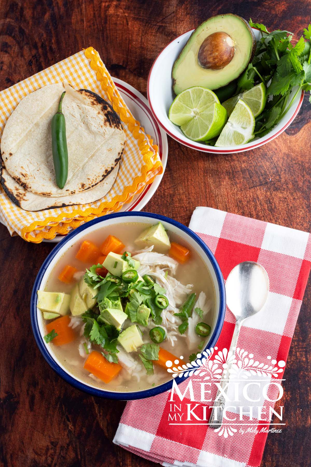 Caldo de Pollo (Mexican chicken soup) served in a white bowl, next to another bowl with avocado, cilantro and limes.