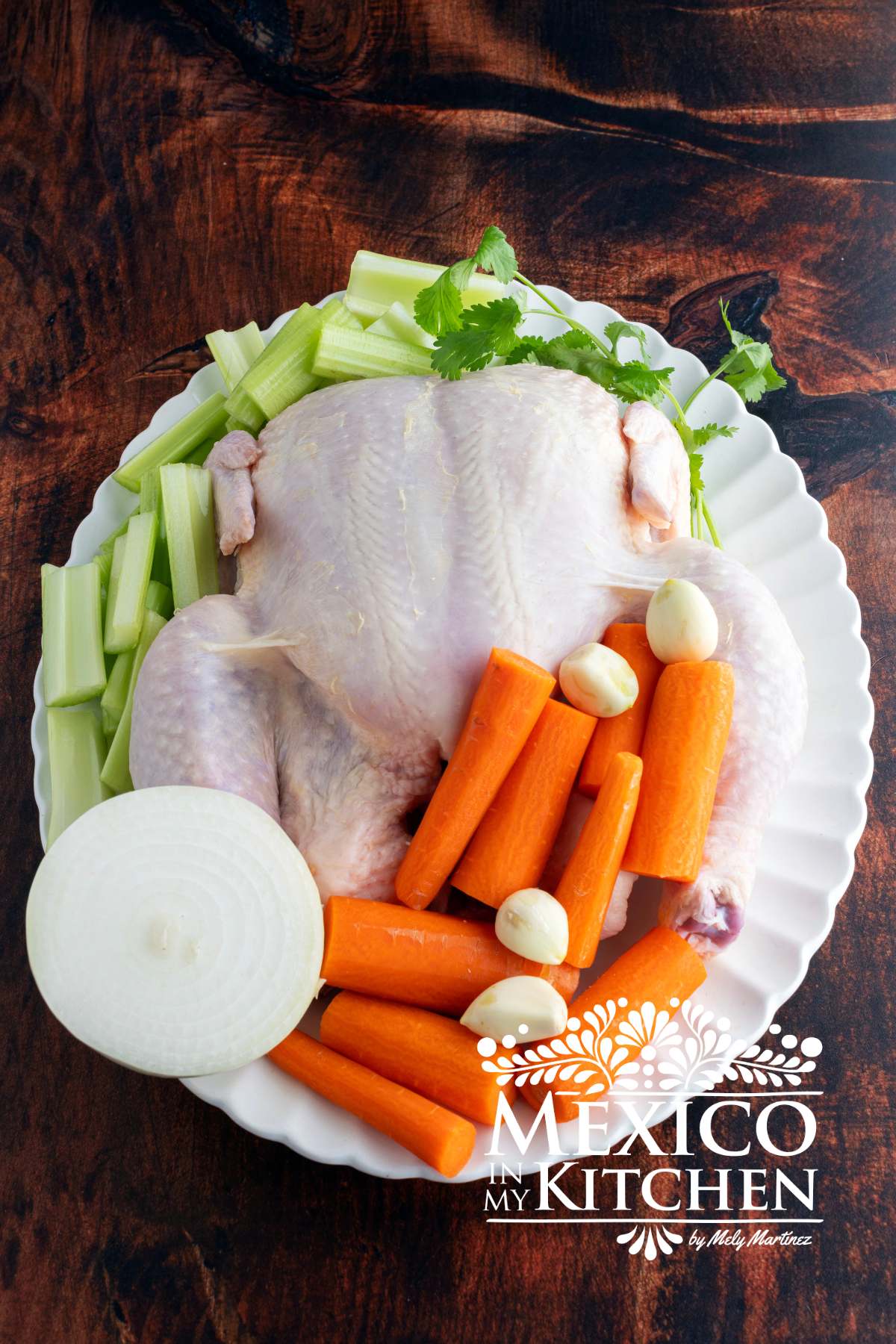 Raw whole chicken, carrots, celery, white onion, cilantro, and garlic cloves served in platter.
