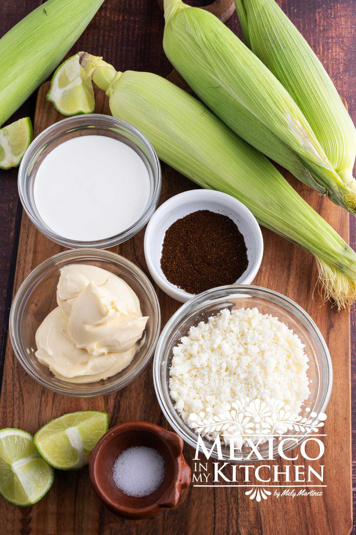Ingredients like corn with husks, Mexican crema, mayo, limes, cotija cheese, and chili powder displays on a cutting board.