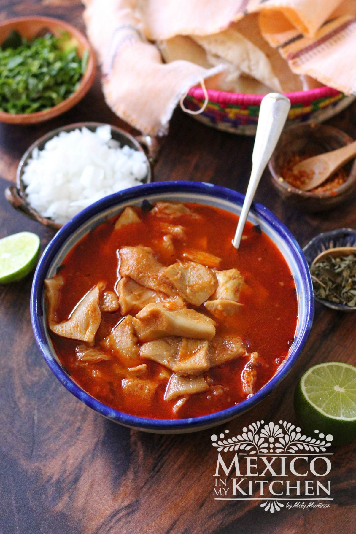 Bowl of Mexican Menudo next to toppings like onion, limes, Mexican oregano and warm tortillas.