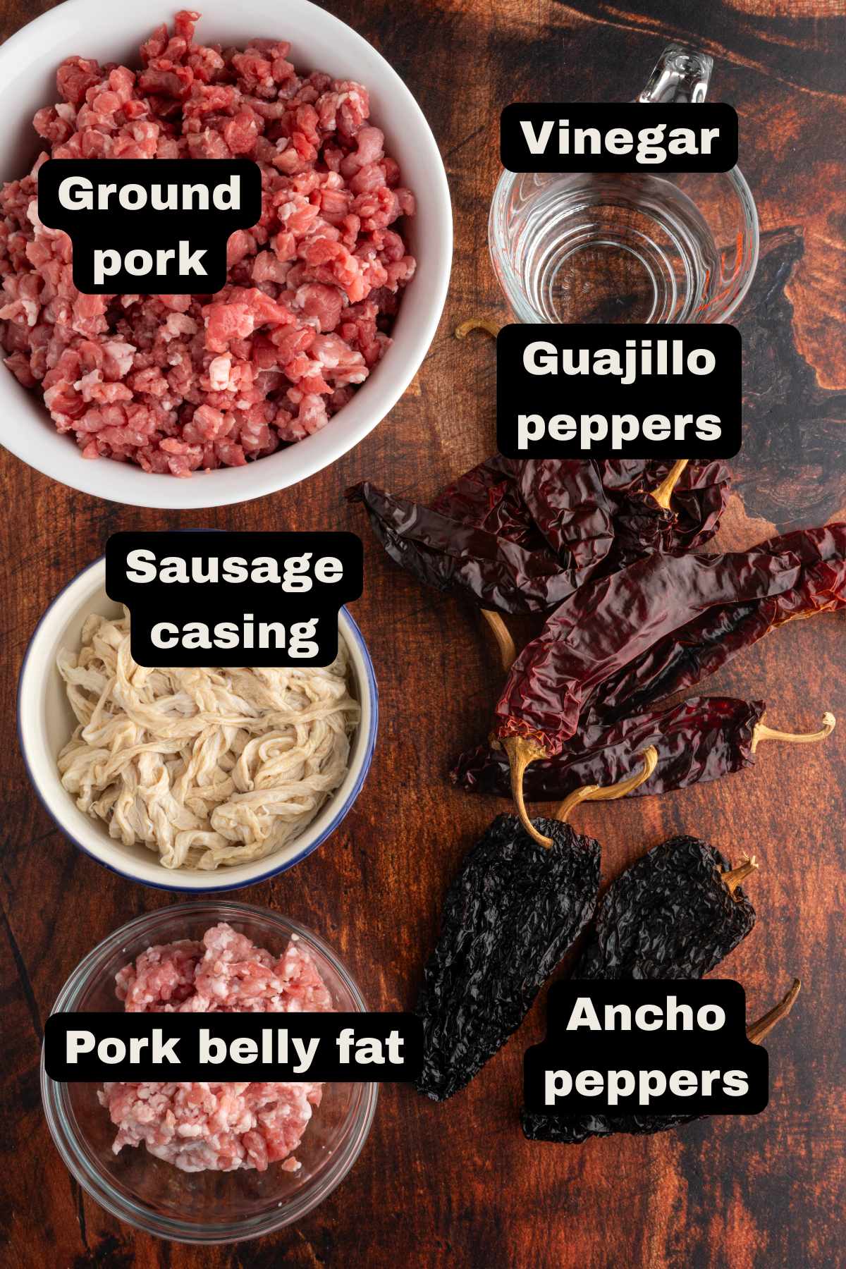 Ground pork, vinegar, guajillo peppers, sausage casing, ancho peppers, and pork belly fat displayed in a table.